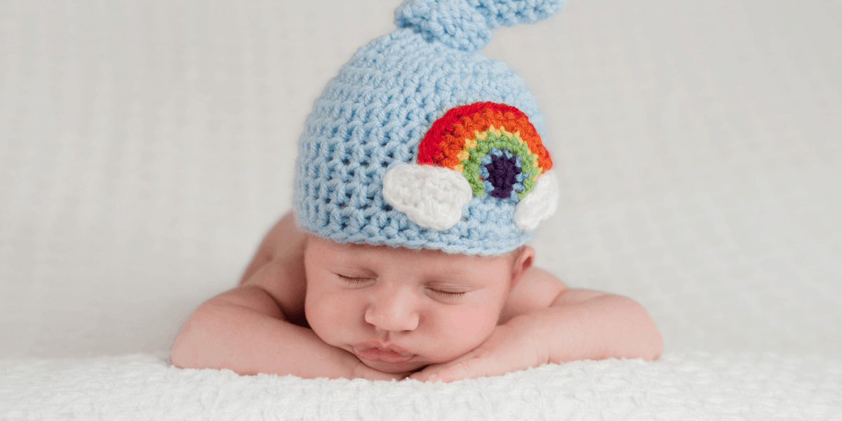 Baby is laying on his tummy, he is asleep and is wearing a blue hat with a rainbow on it.