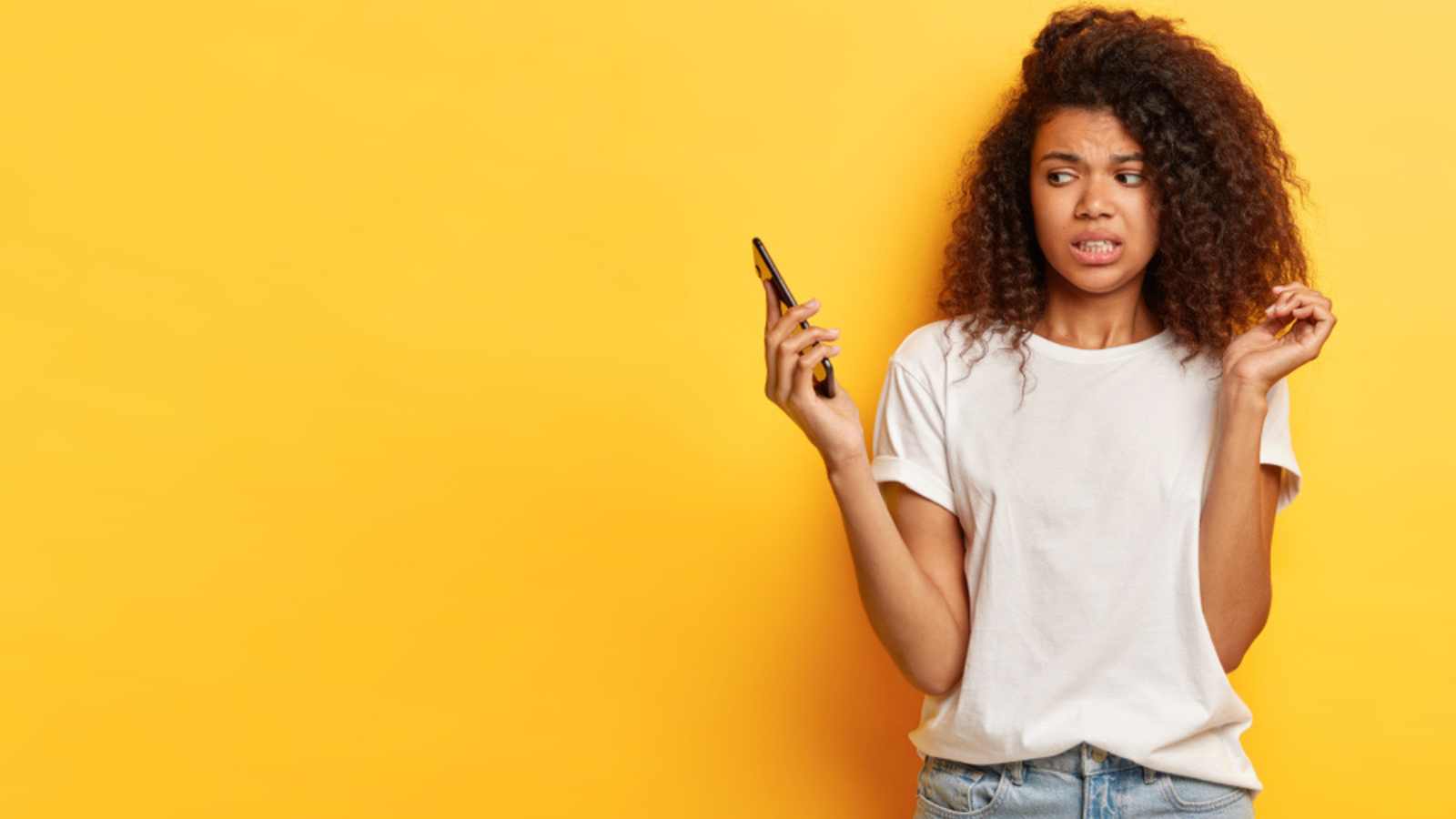 Beautiful female with frizzy Afro hairstyle, looks in displeasure at cell phone